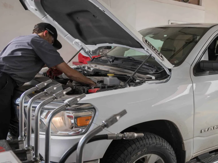 A mechanic performs Battery Services under the hood of a white pickup truck while a person sits in the driver’s seat at an auto repair shop.